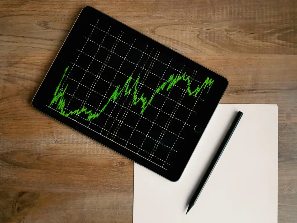 A tablet displaying a green line graph rests on a wooden surface beside a sheet of paper and a pen.